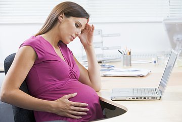 Mutterschaftsgeld in Westfalen-Lippe Das Foto zeigt eine hochschwangere Frau im Büro, die an ihrem Schreibtisch sitzt, vor sich einen Laptop, den Kopf müde mit der linken Hand haltend, während die rechte den Bauch seitlich hält.