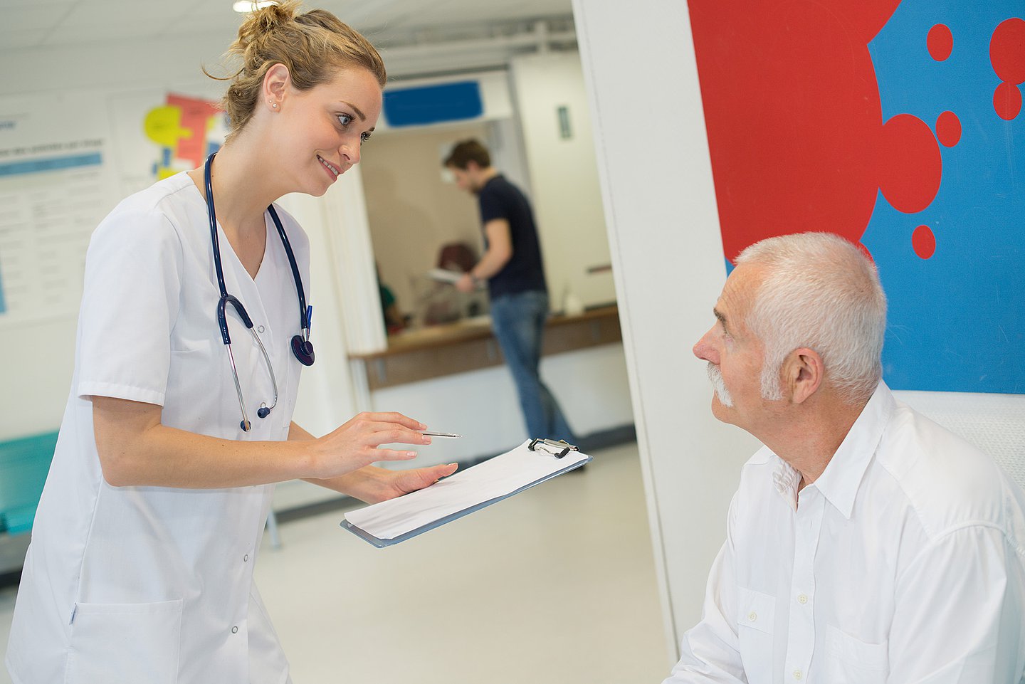 Ein Mann sitzt in der Wartezone in der Notaufnahme eines Krankenhauses und wird von einer Ärztin befragt, die seine Antworten auf ein Dokument einträgt.