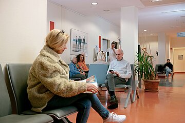 Im Wartezimmer: links vorn unscharf im Bild sitzt eine &auml;ltere Dame. Ihm gegen&uuml;ber sitzt ein Senior und liest Zeitung. Hinten links sitzt ein junger Mann, neben ihm eine alte Dame. Hinten links steht die T&uuml;r offen, ein Arzt ruft einen neuen Patienten auf.