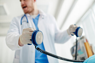 Ein Arzt im wei&szlig;en Kittel und Stethoskop um den Hals h&auml;lt in seinen behandschuhten H&auml;nden die Elektroden f&uuml;r die Reanimation eines nicht sichtbaren Notfall-Patienten in der Hand.