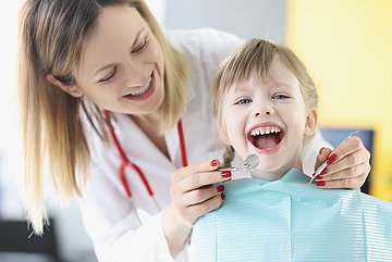 Zahnprophylaxe bei Kindern in Schleswig-Holstein Ein Mädchen sitzt beim Zahnarzt auf dem Behandlungsstuhl, den Mund lachend geöffnet. Eine Mitarbeiterin steht hinter ihr und hält die Instrumente zur Untersuchung bereit.