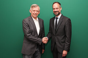 Vor gr&uuml;nem Hintergrund stehen zwei m&auml;nnliche Personen in gesch&auml;ftsm&auml;&szlig;iger Kleidung, die sich die Hand sch&uuml;tteln.