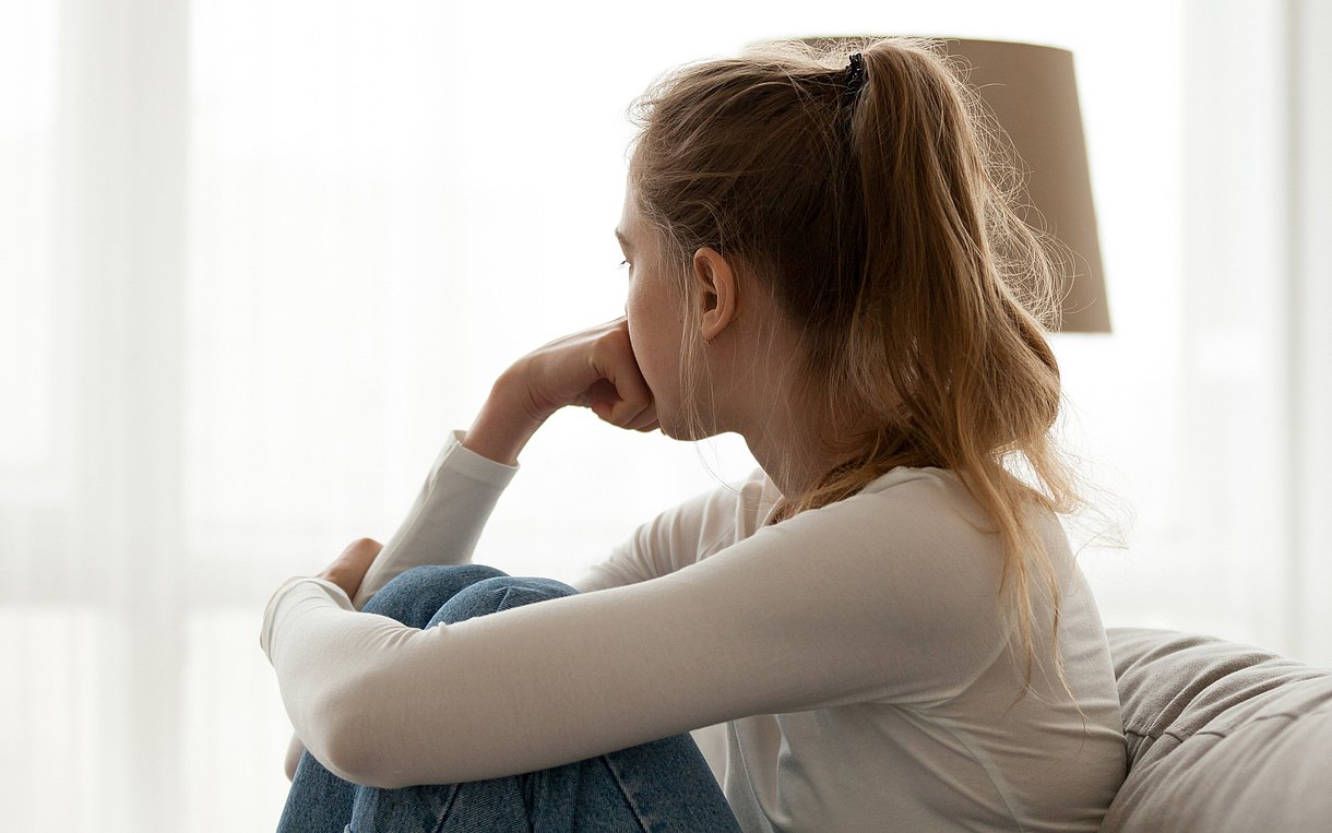 Foto einer jungen Frau von hinten, die nachdenklich aus dem Fenster blickt, während sie auf einem Sofa sitzt