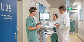 Auf einem Krankenhausgang stehen drei Personen in OP-Kleidung, zwei M&auml;nner und eine Frau, und unterhalten sich. Sie stehen an einem Monitor und haben Unterlagen in der Hand