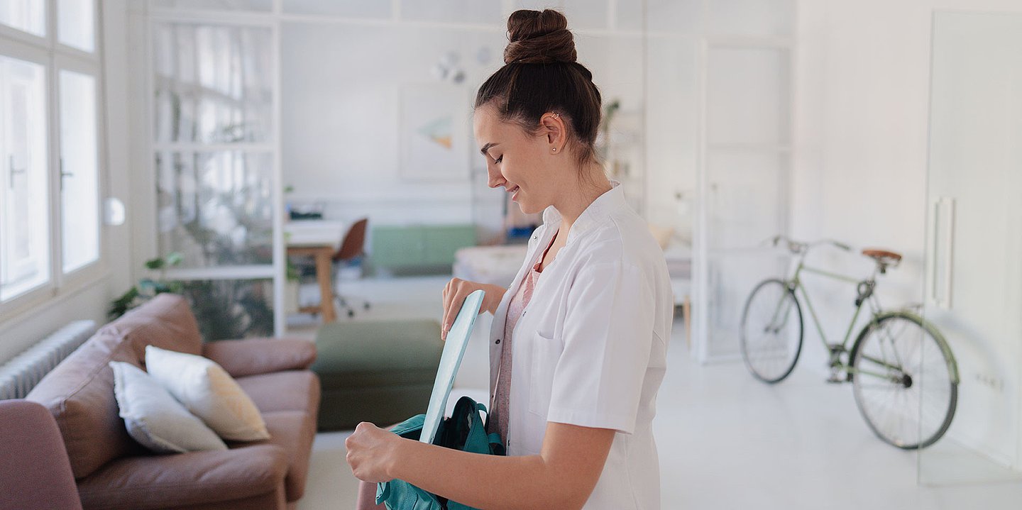 Eine junge Frau im Arztkittel packt in ihrem modernen Wohnzimmer Unterlagen in ihren Rucksack.