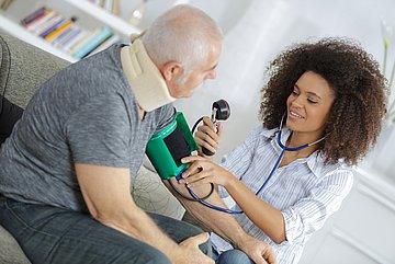 forsa-Umfrage zur Primärversorgung in Schleswig-Holstein Ein älterer Patient mit angelegter Halsmanschette sitzt auf einem Sofa, während eine Ärztin bei ihm den Blutdruck misst.