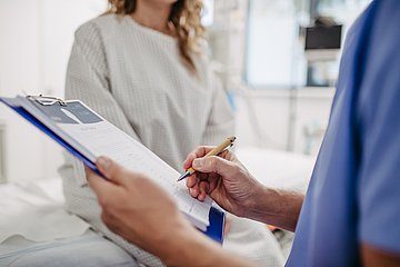 Eine Frau im Patientenkittel sitzt auf der Kante einer Liege im Behandlungsraum eines Krankenhauses und wird von einer Klinikmitarbeiterin befragt, die die Antworten mit einem Stift in ein Formular auf dem Klemmbrett einträgt.
