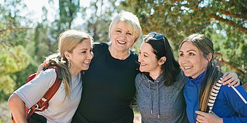Wechseljahre Vier Frauen stehen lachend im Park und legen die Arme umeinander.