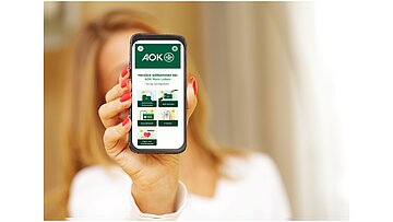 Foto zeigt eine nicht sichtbare Frau im Hintergrund, die das Display ihres Handys in die Kamera zeigt, auf dem die AOK-App 'AOK Mein Leben' u.a. mit der neuen Funktion zur Organspende sichtbar ist..zur 