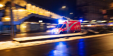 Auf einer Stra&szlig;e bei Nacht f&auml;hrt ein Krankenwagen mit Blaulicht unter einer U-Bahn-Br&uuml;cke durch.