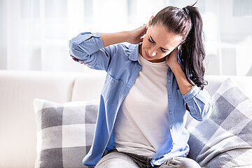 Eine junge Frau sitzt auf dem Sofa und greift sich mit schmerzverzerrtem Gesicht mit beiden H&auml;nden an den Nacken.
