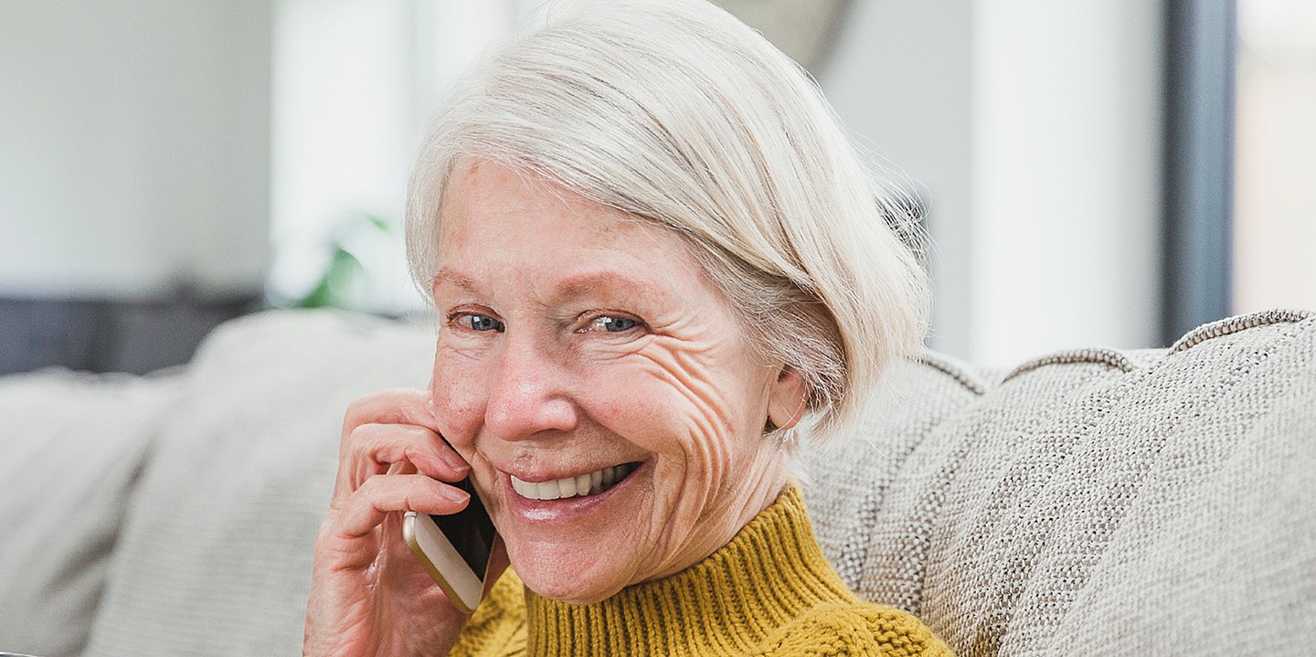 Foto einer älteren Frau, die telefonierend auf einem Sofa sitzt und lacht.