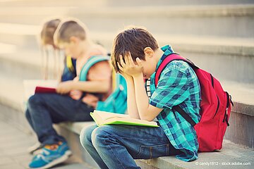 Ein Kind sitzt mit einem Buch auf dem Scho&szlig; auf einer Treppe und vergr&auml;bt den Kopf in seinen H&auml;nden. Im Hintergrund sind zwei weitere Kinder. 