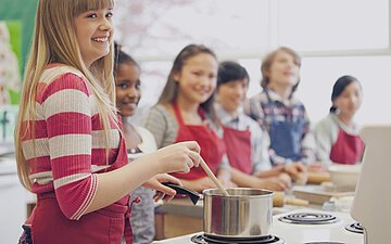 Im Vordergrund steht ein l&auml;chelndes M&auml;dchen mit Sch&uuml;rze und kocht. Im Hintergrund stehen weitere Kinder, die auch kochen.