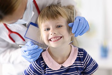 Tag der Zahngesundheit in Schleswig-Holstein Foto zeigt ein lächelndes Kind beim Zahnarztbesuch. Das Mädchen sitzt im Behandlunsgsstuhl, der Zahnarzt steht leicht hinter ihr, hat Mundschutz um und hält mit seinen behandschuhten Händen die Untersuchungsinstrumente nahe am Kopf des Mädchens..