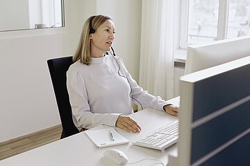 Foto: eine Frau sitzt mit einem Headset in einem B&uuml;ro vor einem Bildschirm 