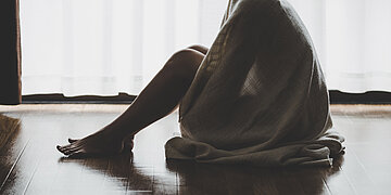 Foto: Eine Frau sitzt auf dem Boden, die Knie angezogen und ein großes Tuch um Schultern und Rücken.