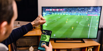 Foto: Ein Mann sitzt mit seinem Smartphone, auf dem eine App f&uuml;r Sportwetten zu sehen ist, vor dem TV-Bildschirm, auf dem ein Fu&szlig;ballspiel l&auml;uft.