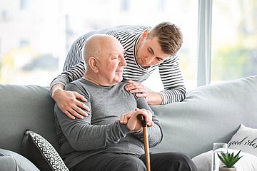 Bild_PM_Pflegende Angehörige_AOK Bayern bietet Workshop-Reihe an Ein junger Mann legt die Hände über die Schultern eines älteren Mannes, der mit einem Gehstock auf einem Sofa sitzt.