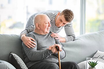 Ein junger Mann legt die H&auml;nde &uuml;ber die Schultern eines &auml;lteren Mannes, der mit einem Gehstock auf einem Sofa sitzt.