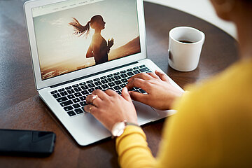 Eine Person sitzt mit dem Laptop irgendwo an einem runden Tisch und recherchiert im Internet. Auf dem Bildschirm ist die obere H&auml;lfte einer laufenden Person zu sehen, im Hintergrund die auf- oder untergehende Sonne.