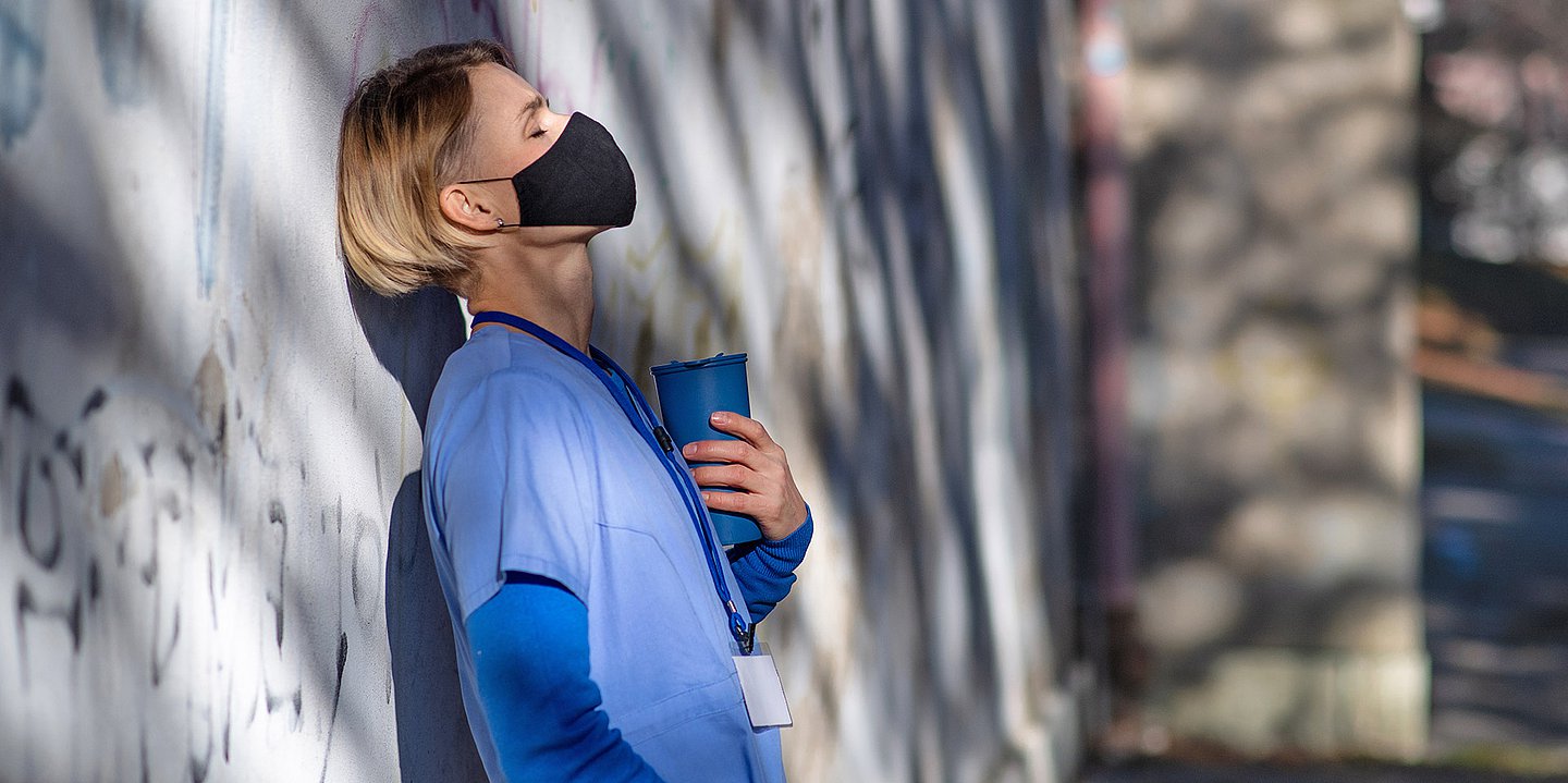 Foto einer erschöpften Pflegerin mit Maske, die sich in blauer Arbeitskleidung draußen an eine Wand lehnt