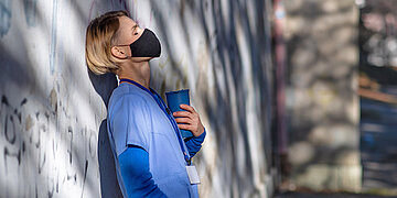 Foto einer ersch&ouml;pften Pflegerin mit Maske, die sich in blauer Arbeitskleidung drau&szlig;en an eine Wand lehnt
