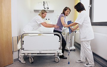 Frau mit Schmerzen versucht aus ihrem Krankenhausbett aufzustehen. Zwei Krankenschwestern helfen ihr.