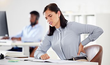Rückenschmerzen Foto: Eine Frau sitzt am Schreibtisch und fasst mit der Hand an ihren schmerzenden Rücken.