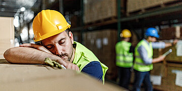 In einer Lagerhalle lehnt sich ein Bauarbeiter an ein Regal mit Holz. Er hat die Augen zu und es sieht aus, als w&uuml;rde er schlafen. Im Hintergrund arbeiten zwei seiner Kollegen.
