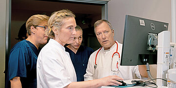 Mehrere medizinische Fachkr&auml;fte stehen vor einem Computer und besprechen gemeinsam die Patientendaten auf dem Bildschirm.
