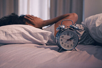 Eine Frau in Nahaufnahme liegt im Bett und schl&auml;gt die H&auml;nde vors Gesicht. Der Wecker zeigt auf 2:15 Uhr.
