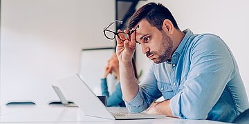 Foto: Ein Mann sitzt am Laptop, die Brille in der Hand, und schaut auf den Bildschirm.