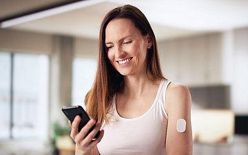AOK Mein Leben-App Bild einer Frau mittleren Alters in sommerlichem Outfit, die lächelnd in ihrem Wohnzimmer auf ihr Smartphone blickt