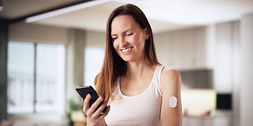 AOK Mein Leben-App Bild einer Frau mittleren Alters in sommerlichem Outfit, die lächelnd in ihrem Wohnzimmer auf ihr Smartphone blickt