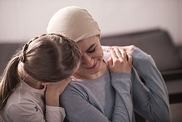 Ambulante Hospizdienste in Westfalen-Lippe Foto zeigt eine kranke junge Frau auf einem Sofa sitzend, die sich an eine sich ihr zuwendende Frau anlehnt, die sie an den Schultern hält.