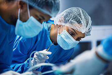 Zwei Operateure in OP-Kleidung beugen sich &uuml;ber den OP-Tisch, die &Auml;rztin h&auml;lt ein Instrument in der Hand.