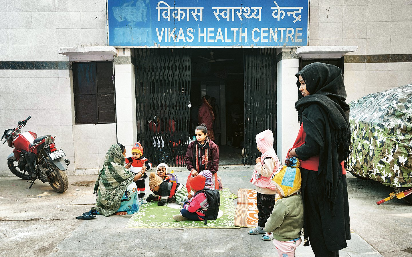Auf einem Teppich vor einem Gebäude des Vikas Health Centre sitzen mehrere Mütter mit ihren Kindern. 