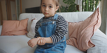 Ein kleines M&auml;dchen mit Zopf und blauer Jeans-Latzhose sitzt mit traurigem Gesicht auf einem wei&szlig;en Sofa und h&auml;lt sich mit der rechten Hand das linke Handgelenk
