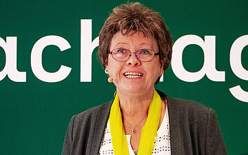 Auf dem Podium steht Angela Clausen und h&auml;lt l&auml;chelnd einen Vortrag. Hinter ihr steht eine Wand mit der Aufschrift "Selbsthilfe-Fachtagung".
