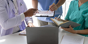Foto: Mehrere Pflegekr&auml;fte stehen um einen Laptop herum, eine Person h&auml;lt eine Liste aus Papier.