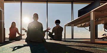 Drei Personen meditieren im Sonnenlicht vor einem großen Fenster.