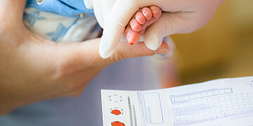 Eine Hand nimmt einen kleinen Bluttropfen von der Ferse eines Neugeborenen.