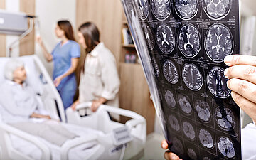 Foto: Ein Arzt h&auml;lt CT-Aufnahmen von Gehirn hoch, im Hintergrund liegt eine Patientin im Krankenhausbett.