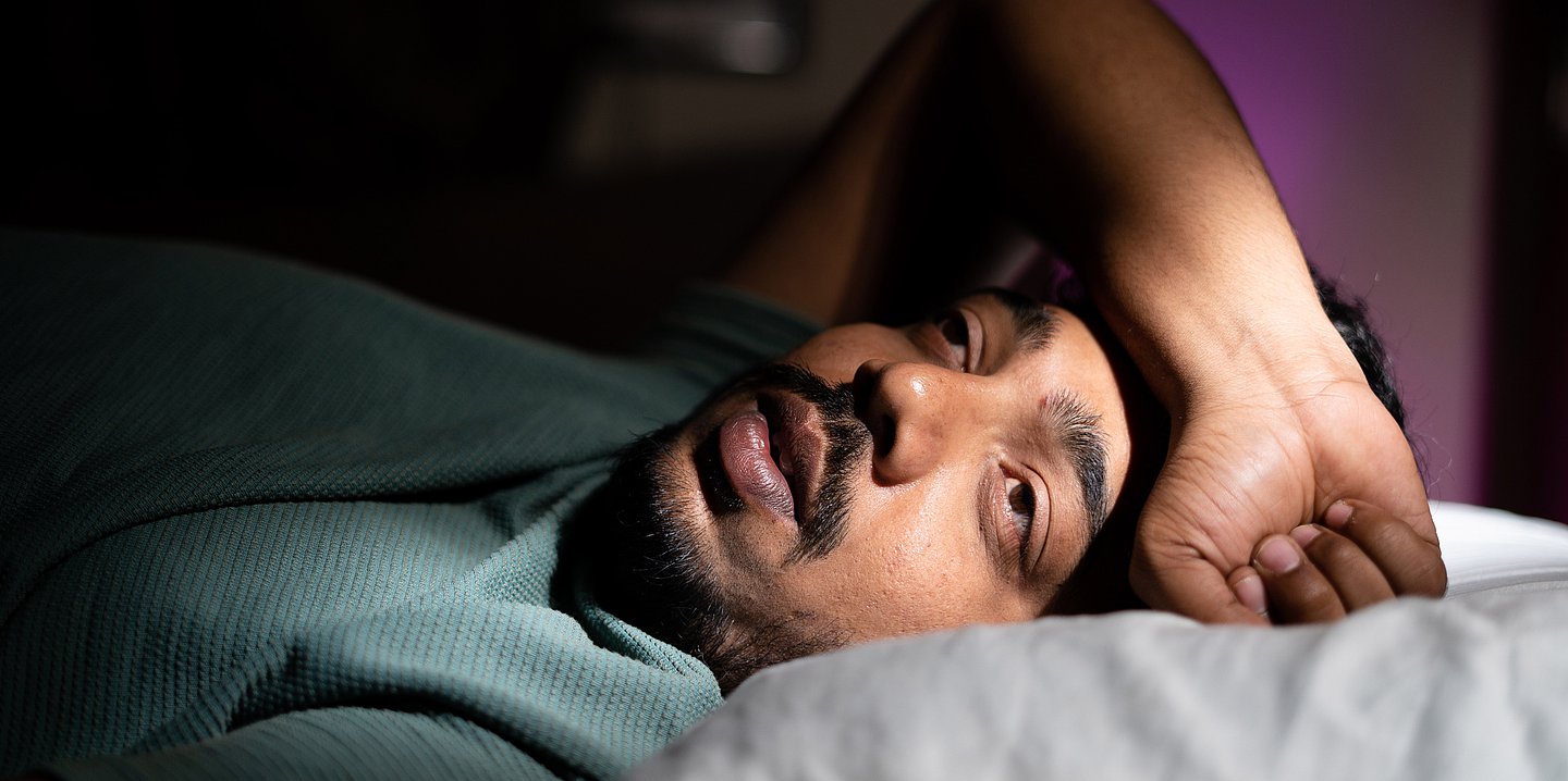Foto: Ein Mann liegt mit offenen Augen in seinem Bett, den Arm hat er über der Stirn abgelegt. Er sieht sehr müde und erschöpft aus.