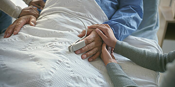Bild: Eine Person liegt in einem Krankenbett, eine andere Person h&auml;lt seine rechte Hand in den ihren.