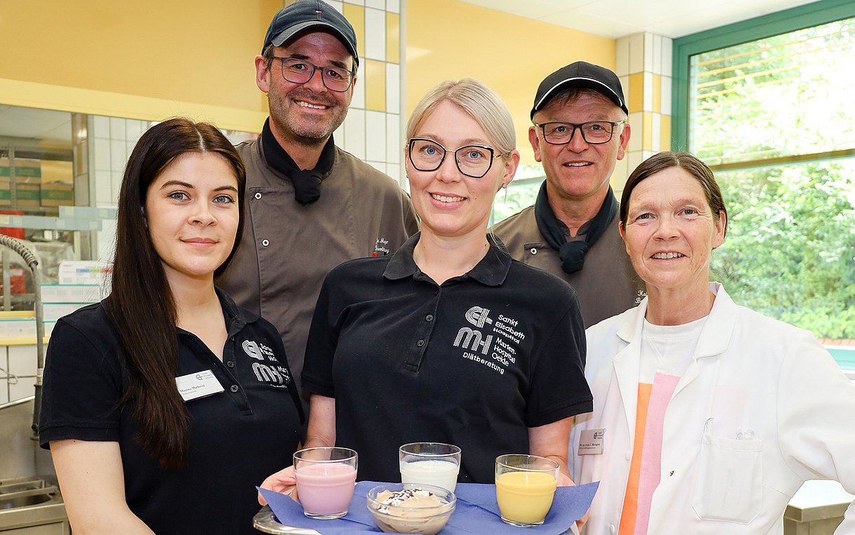 Foto: Ernährungsteam des Gütersloher Sankt Elisabeth Hospital, fünf Personen, davon zwei Männer und drei Frauen stehen zusammen und lächeln in die Kamera. Eine Frau trägt ein Tablett mit Trinken und Essen.