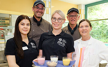 Foto: Ern&auml;hrungsteam des G&uuml;tersloher Sankt Elisabeth Hospital, f&uuml;nf Personen, davon zwei M&auml;nner und drei Frauen stehen zusammen und l&auml;cheln in die Kamera. Eine Frau tr&auml;gt ein Tablett mit Trinken und Essen.