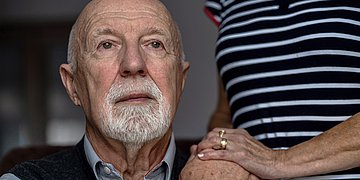 Alter Mann schaut in die Ferne, seine Frau hat die Hand auf seiner Schulter. Der alte Mann hält diese Hand.