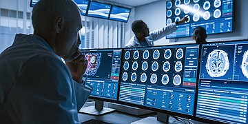 Ein Arzt sitzt am Schreibtisch vor 3 Monitoren. Auf ihnen zu sehen sind verschiedene Hirnscans.